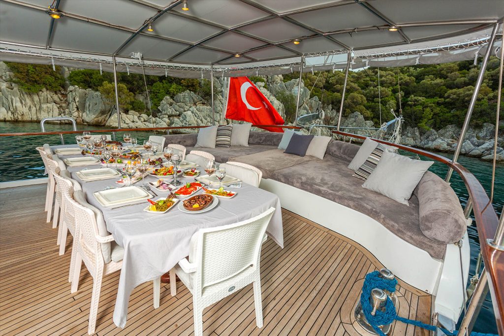 A covered yacht deck with a table set for a meal, featuring white chairs and a variety of dishes. A large cushioned lounge area and a Turkish flag are visible, with water and rocky, tree-covered shore in the background.