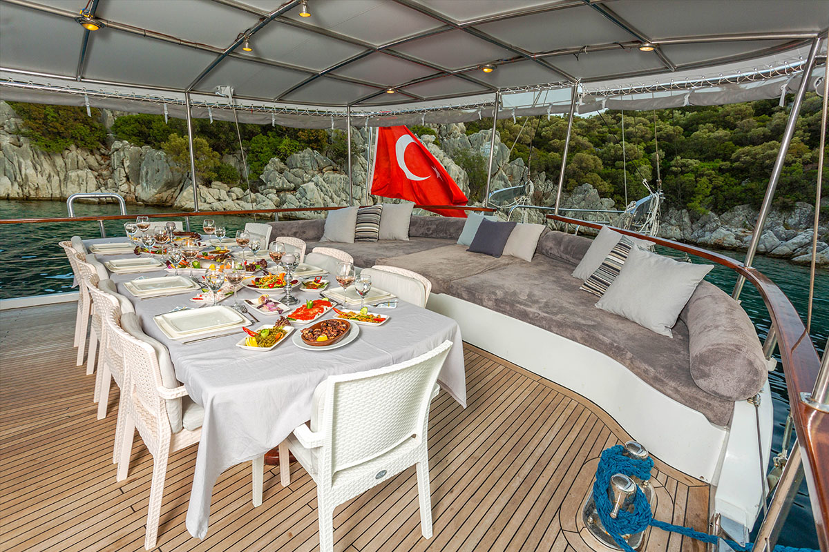 A covered yacht deck with a table set for a meal, featuring white chairs and a variety of dishes. A large cushioned lounge area and a Turkish flag are visible, with water and rocky, tree-covered shore in the background.