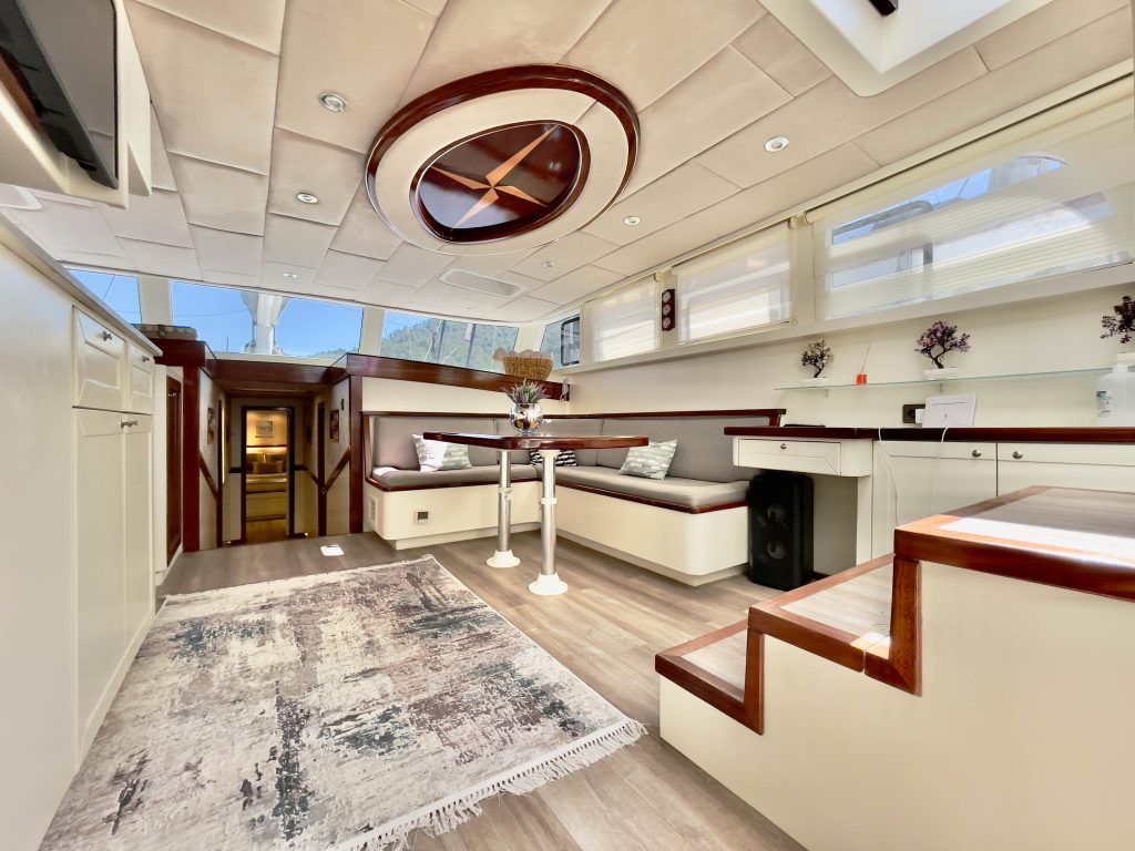 Spacious, modern yacht interior featuring white walls, wood accents, a seating area with cushions, dining table, steps, and large windows allowing natural light to fill the room. A patterned rug decorates the floor.