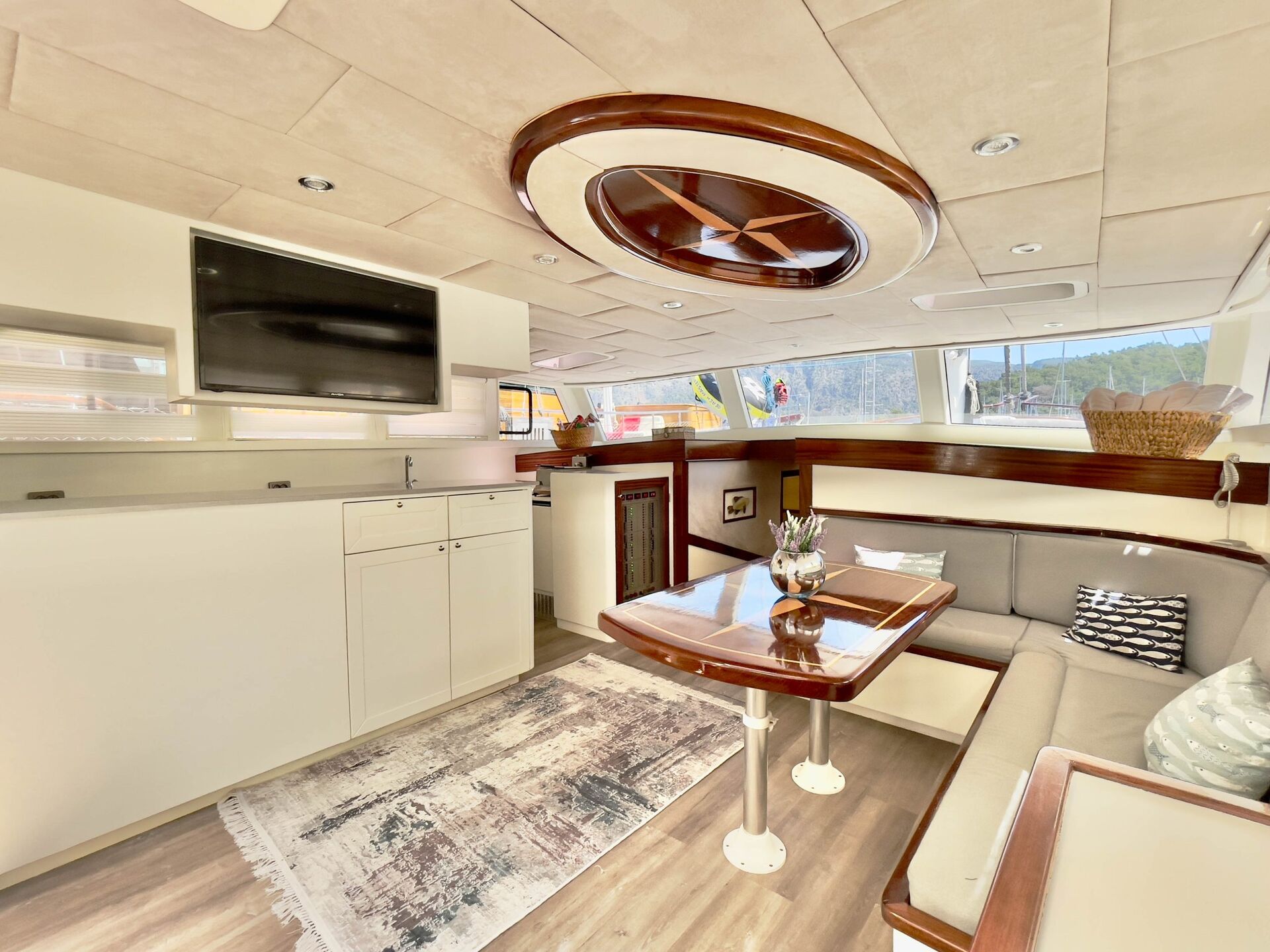 Bright, modern yacht interior with cream-colored walls, an L-shaped sofa, a wooden table, a TV mounted on the wall, and large windows letting in natural light. A rug and decorative pillows add comfort to the space.