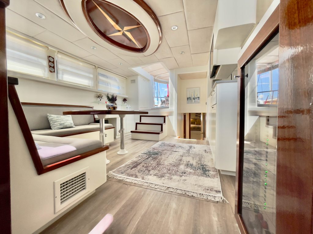 A bright, modern yacht interior with wood accents, a cozy seating area, dining table, soft rug, and stairs leading to an upper deck. Large windows let in natural light, creating an open, airy atmosphere.