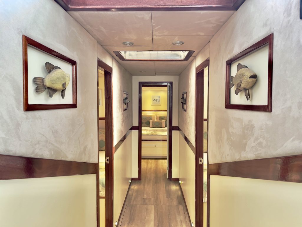 A hallway with wood flooring and cream-colored walls, decorated with framed fish sculptures, leading to a well-lit bedroom with a neatly made bed.