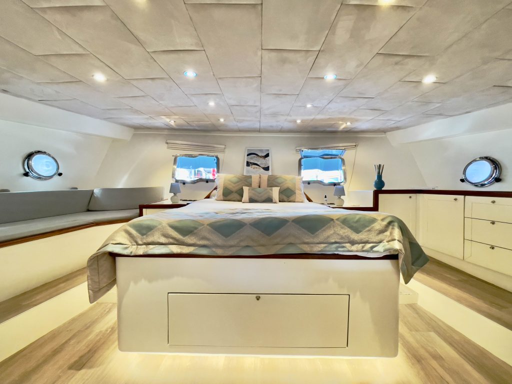 A spacious yacht bedroom with a large bed, patterned bedding, built-in cabinets, round windows, and soft lighting. Decorative pillows and a vase accent the cozy, modern space.