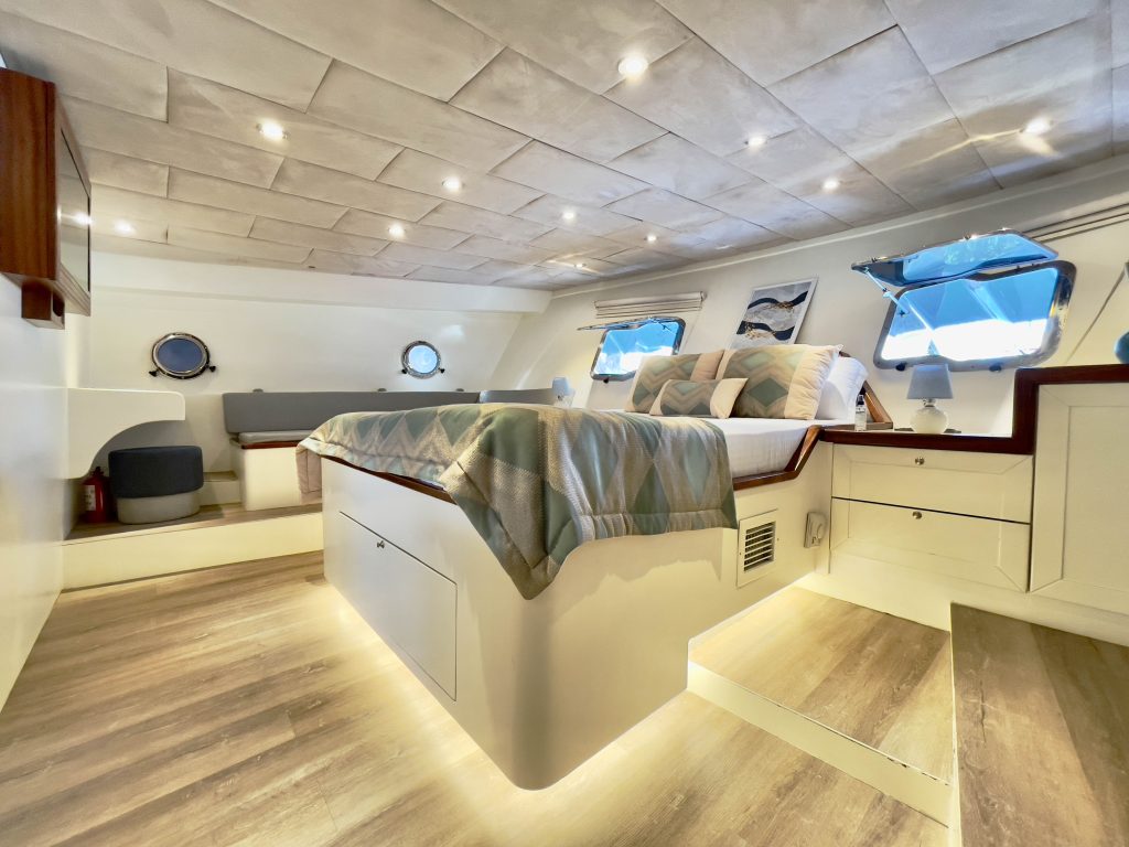 A modern, well-lit yacht cabin with a double bed, patterned bedding, large windows, light wood floors, built-in storage, and soft recessed lighting under the bed and ceiling.