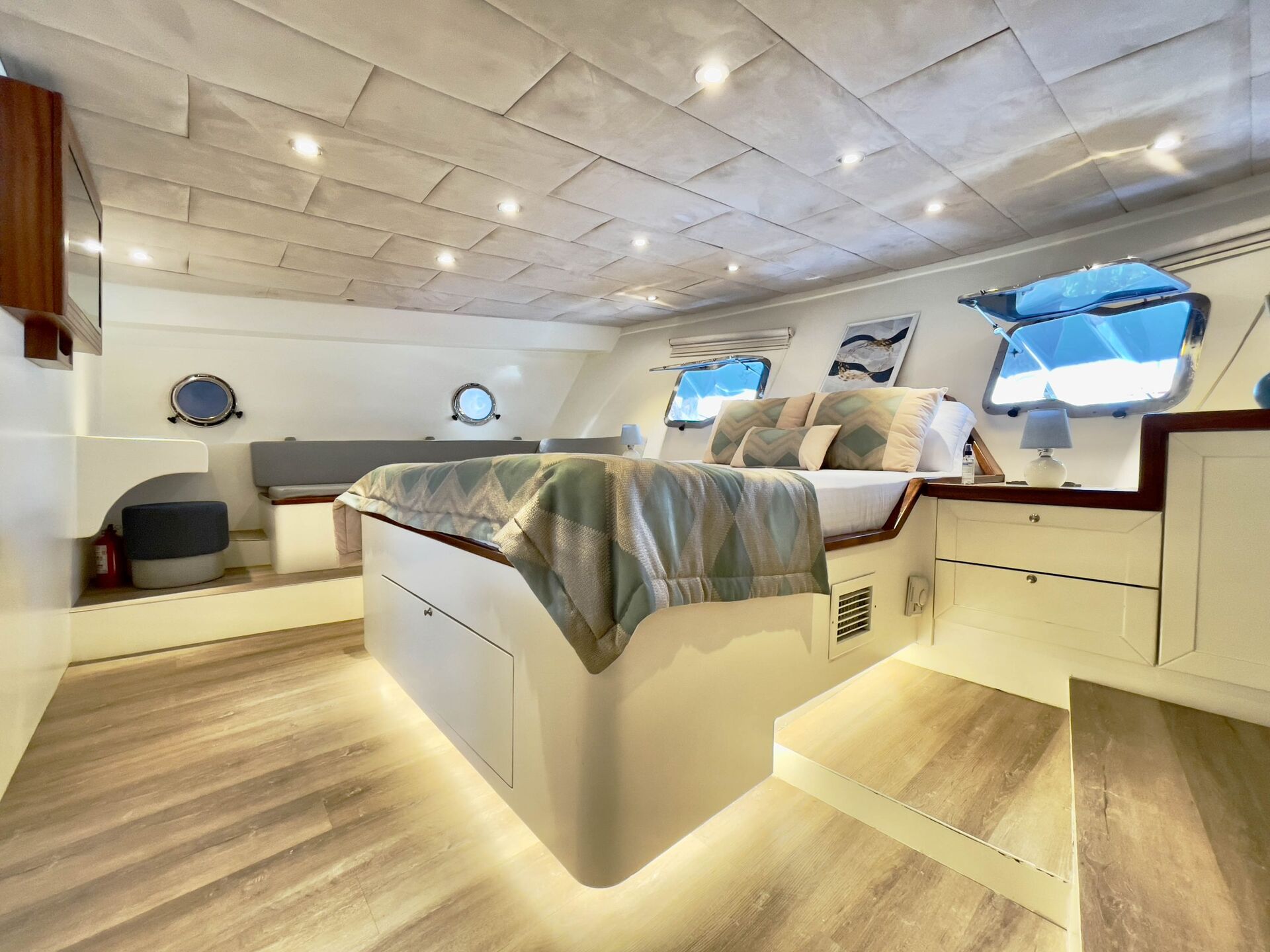 A modern, well-lit yacht cabin with a double bed, patterned bedding, large windows, light wood floors, built-in storage, and soft recessed lighting under the bed and ceiling.