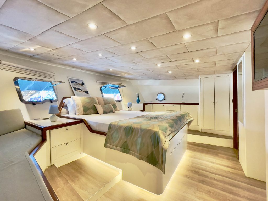 A spacious yacht bedroom with a large bed, patterned bedding, two windows, white cabinets, wood accents, soft lighting, and modern decor, including artwork, lamps, and a vase on a side dresser.