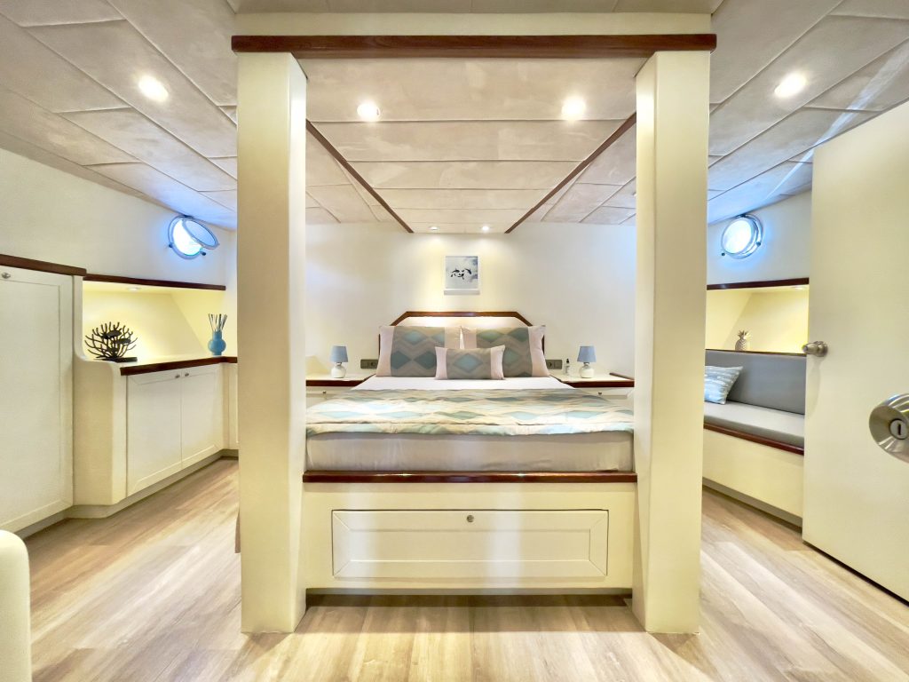 A bright, modern bedroom with a large bed centered under a canopy frame, surrounded by white walls, wood accents, built-in shelves, and small round windows, creating a cozy, nautical atmosphere.
