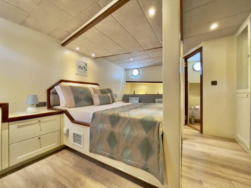 A cozy, modern bedroom with a neatly made bed featuring patterned bedding, bedside tables with lamps, light wood flooring, recessed ceiling lights, and a small round window on the wall. An open door leads to another room.