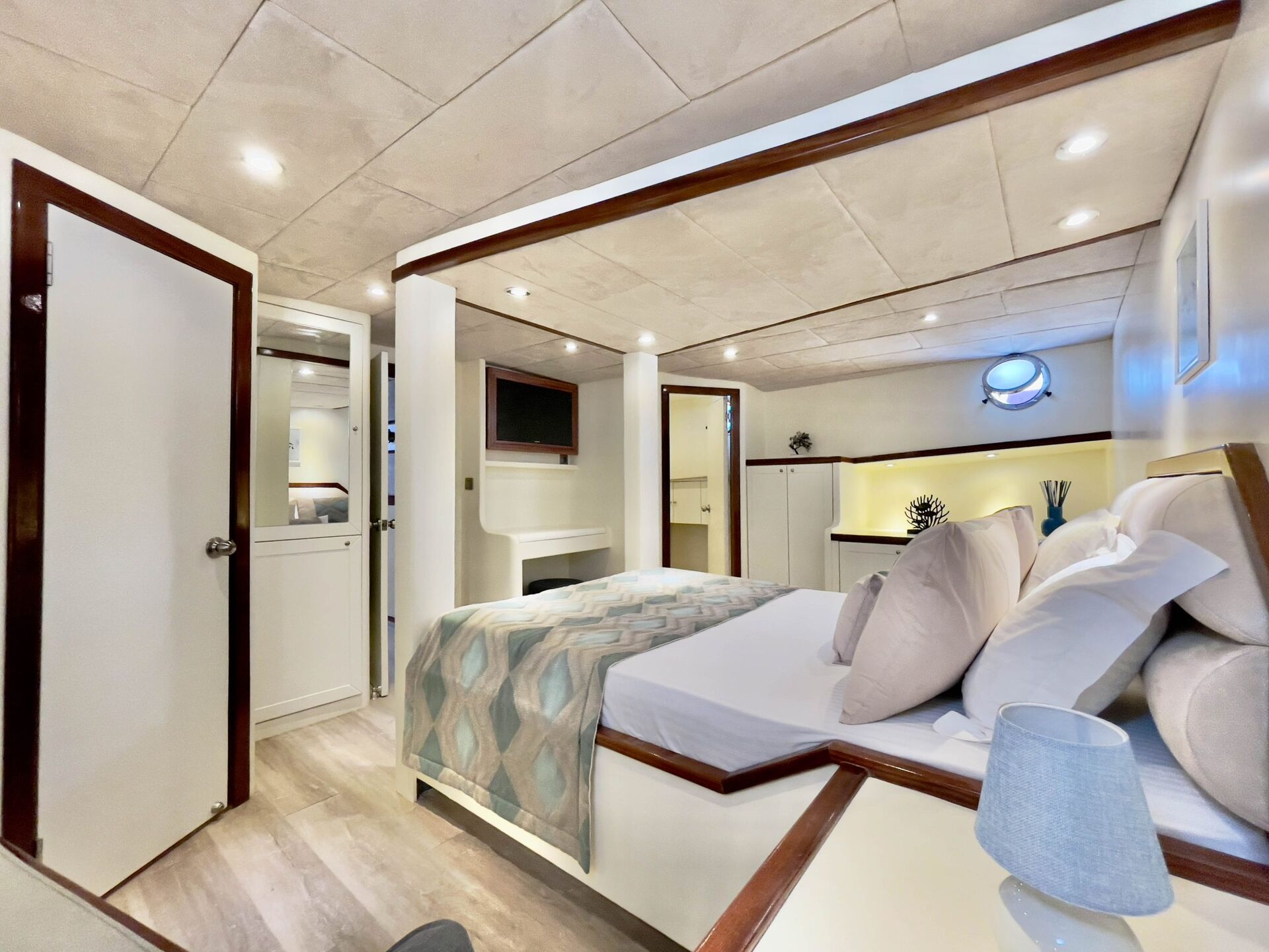 A modern, cozy bedroom with a neatly made bed, patterned blanket, soft pillows, and light wood accents. The room features a small lamp, wall-mounted TV, cabinets, and round porthole window, giving it a nautical, yacht-like feel.