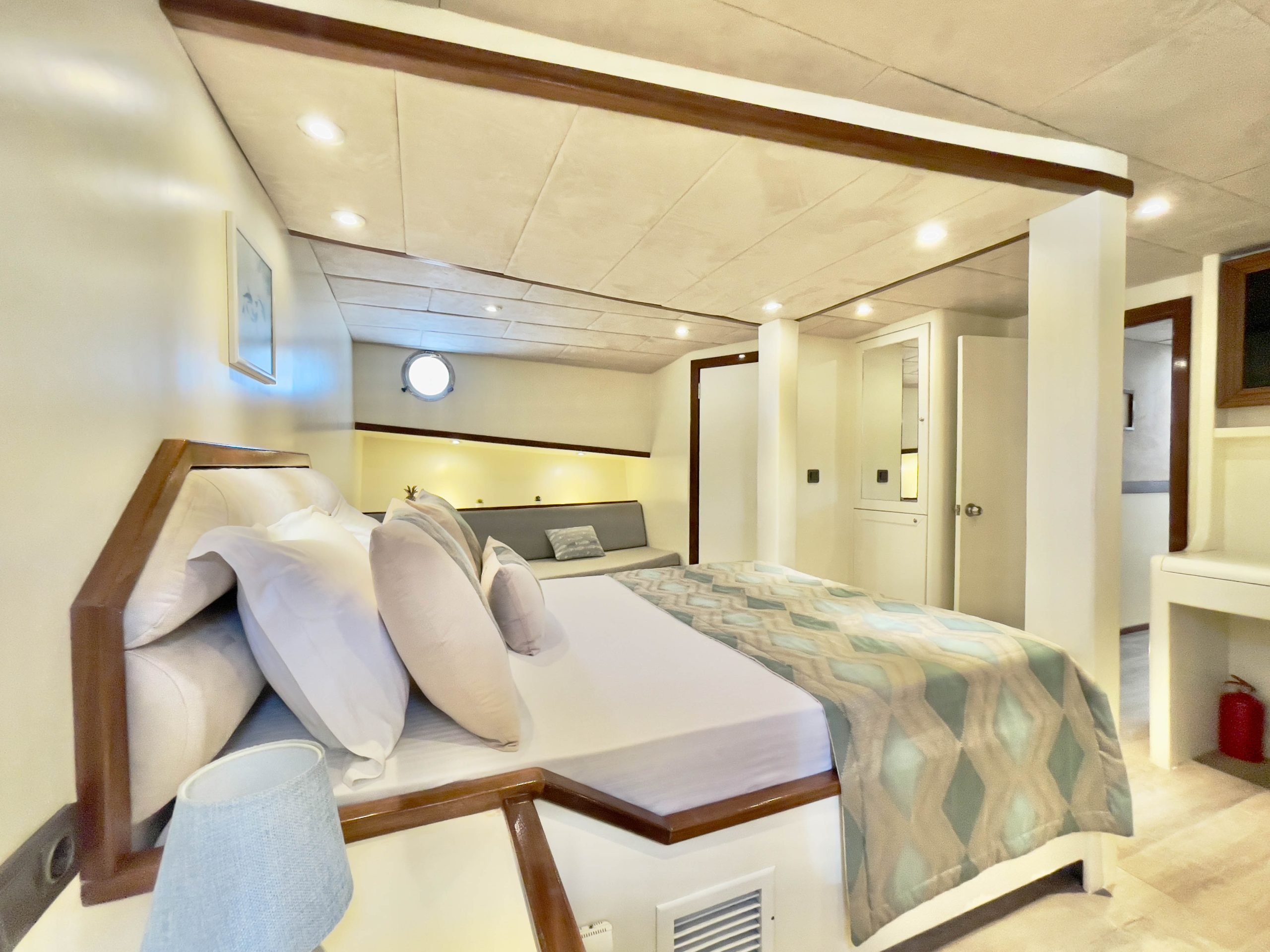 A modern, well-lit bedroom with a large bed, white linens, decorative pillows, a patterned throw, a gray sofa, built-in lighting, and a small round window above the bed. The walls and ceiling are light-colored.