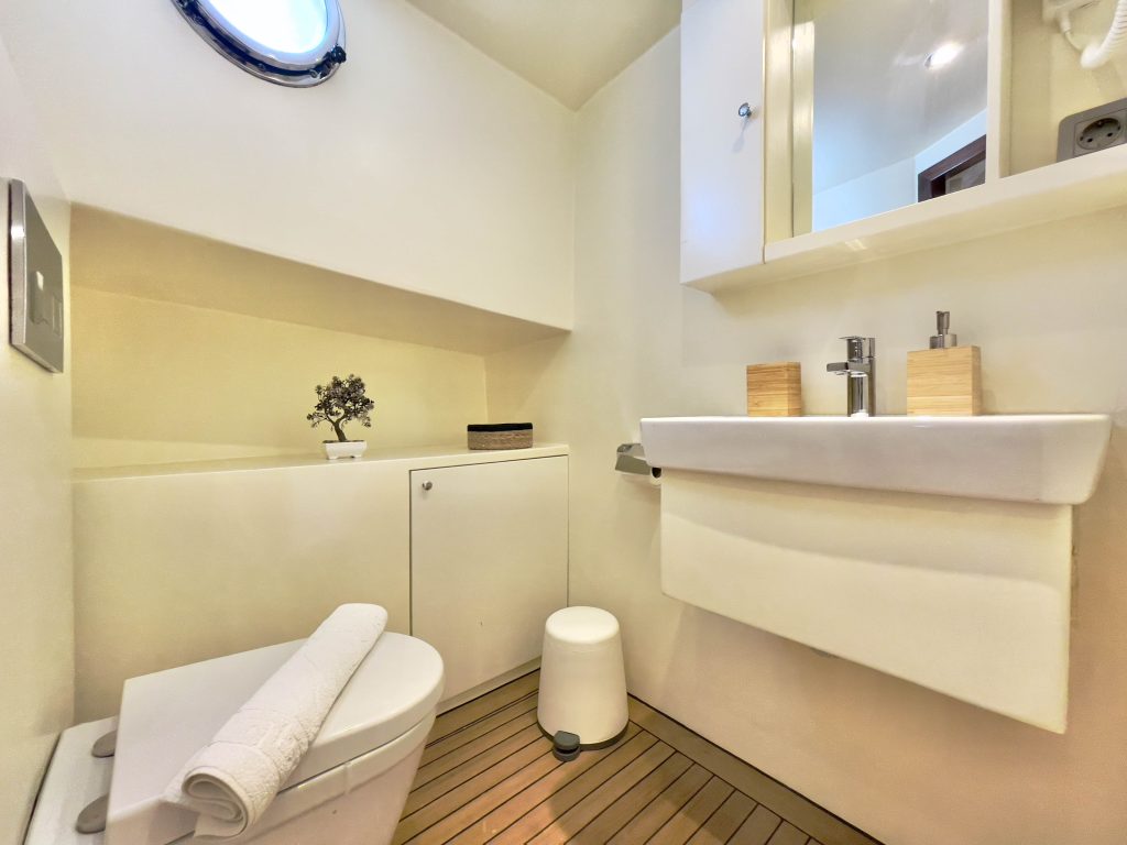 A modern, compact bathroom with a white toilet, rolled towel, rectangular sink, wooden floor, small porthole window, mirror cabinet, and minimalist decor including a plant and soap dispensers.
