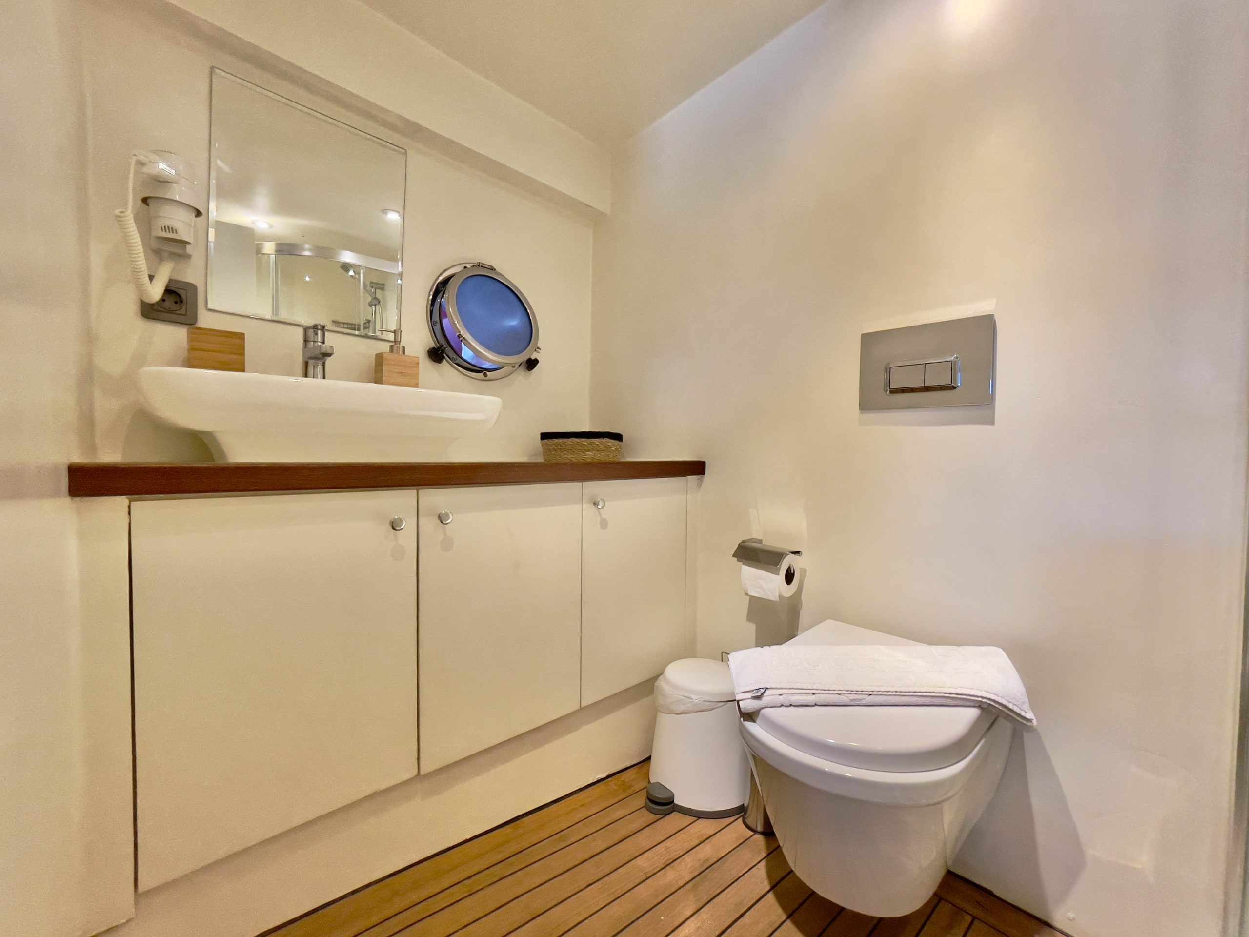 Modern bathroom with white walls, a floating toilet, a towel on the toilet lid, a white sink on a wooden countertop, and cabinets below. There is a circular mirror, a wall-mounted hairdryer, and a wood-style floor.