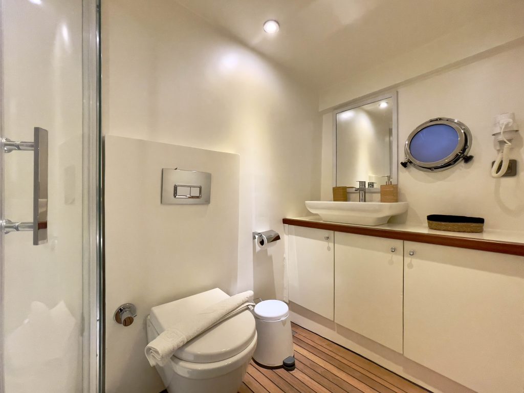 A modern bathroom with a white toilet, sink, and cabinets, wooden floor, a round porthole window, a mirror, and a towel on the toilet lid. The space is bright and neatly organized.