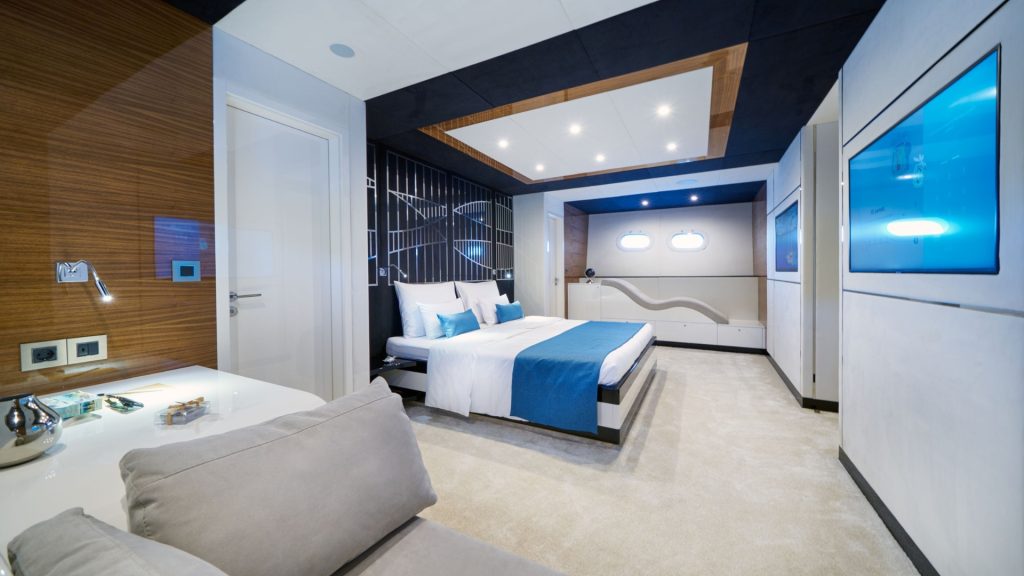 Modern bedroom with a large bed featuring white and blue bedding, sleek furniture, a wall-mounted TV, a desk with a chair, and porthole windows letting in natural light. The room has contemporary decor and carpeting.