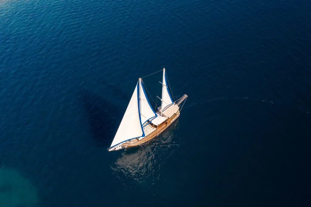 Aerial view of a sailboat with white sails gliding on calm, deep blue water, leaving a gentle wake behind.