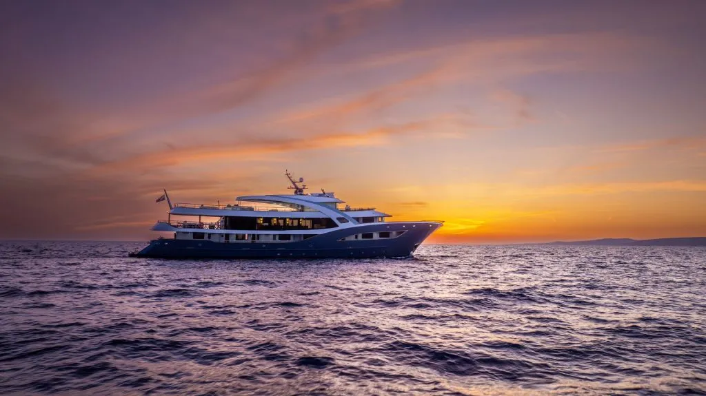 A large yacht sails on calm water at sunset, with the sky glowing in orange and purple hues. The yacht is silhouetted against the horizon, creating a tranquil and luxurious scene.
