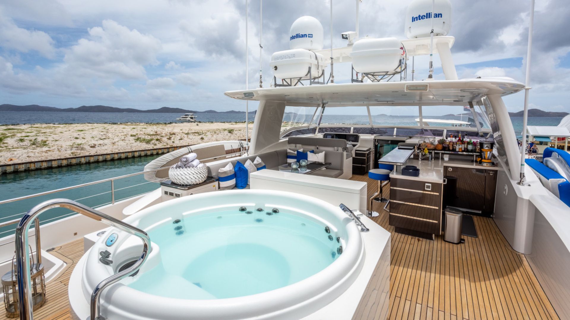 A luxurious yacht deck with a circular hot tub, bar area, sun loungers, and high-tech satellite equipment, docked near a scenic shoreline with hills under a cloudy sky.