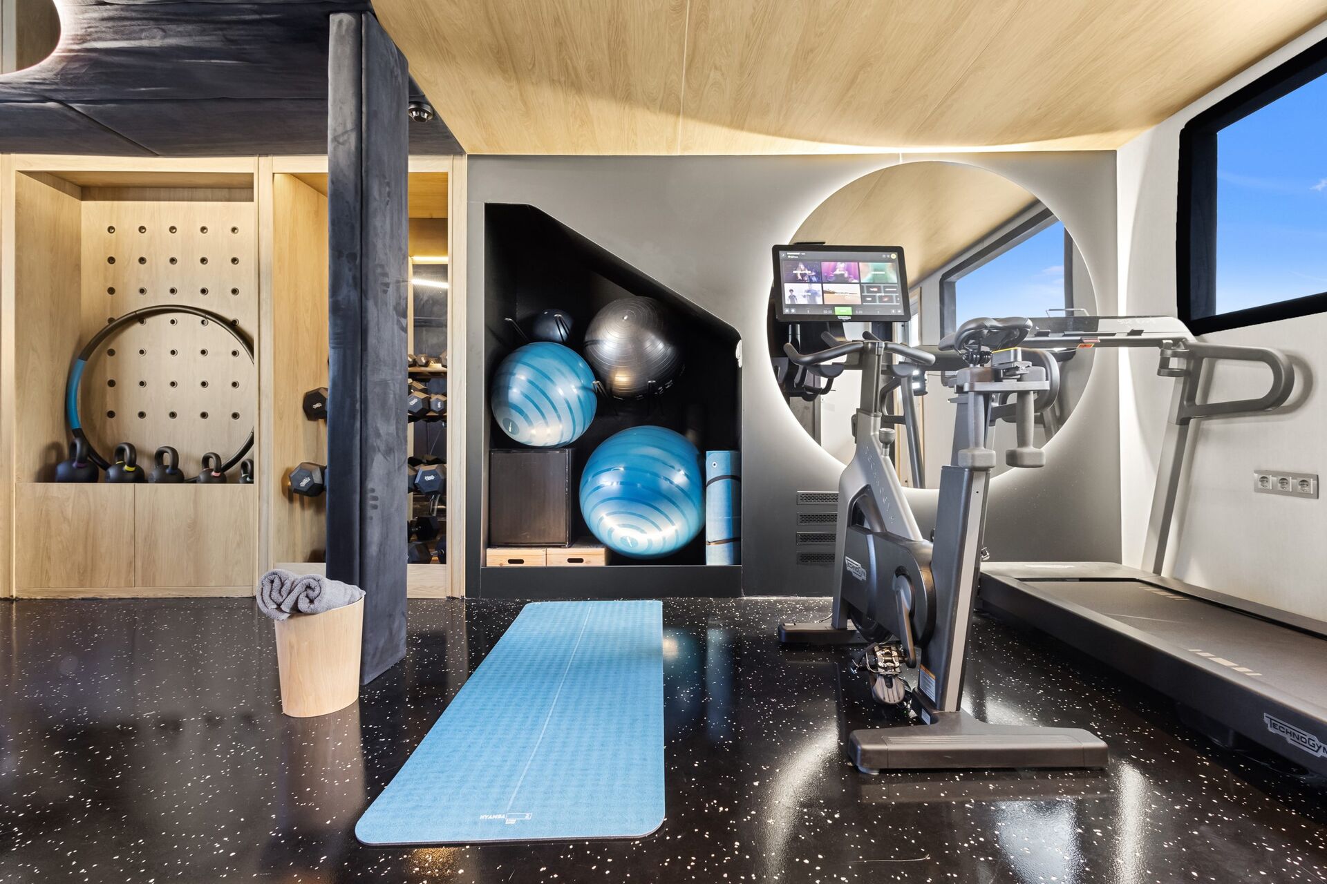 Modern home gym with black speckled flooring, a treadmill, exercise bike, blue yoga mat, resistance balls, weights, and fitness equipment organized on wooden shelves, with a large window providing natural light.