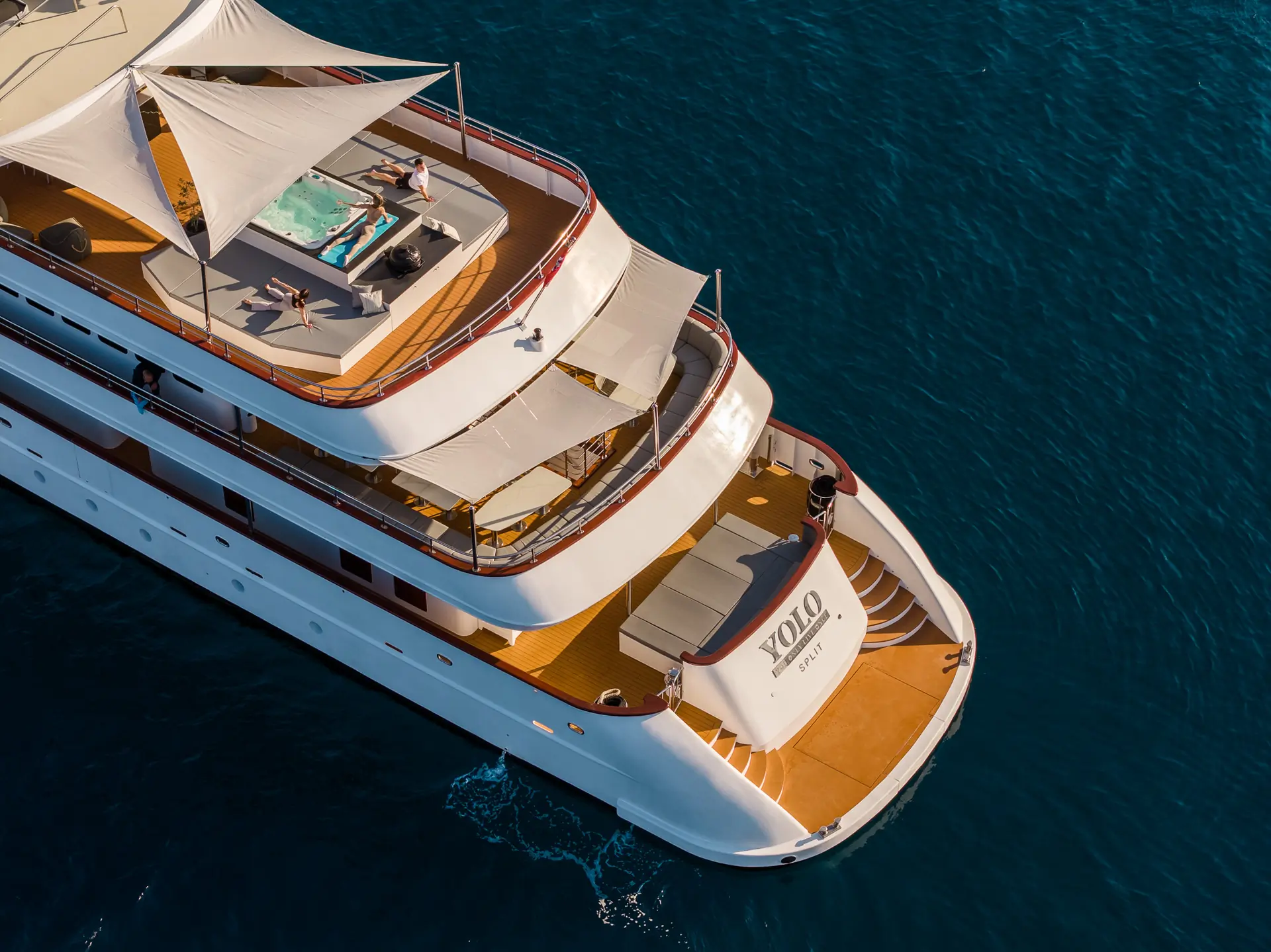 Aerial view of a luxury yacht named YOLO cruising on deep blue water, with people relaxing on a sun deck and near a small pool under white canopy shades.