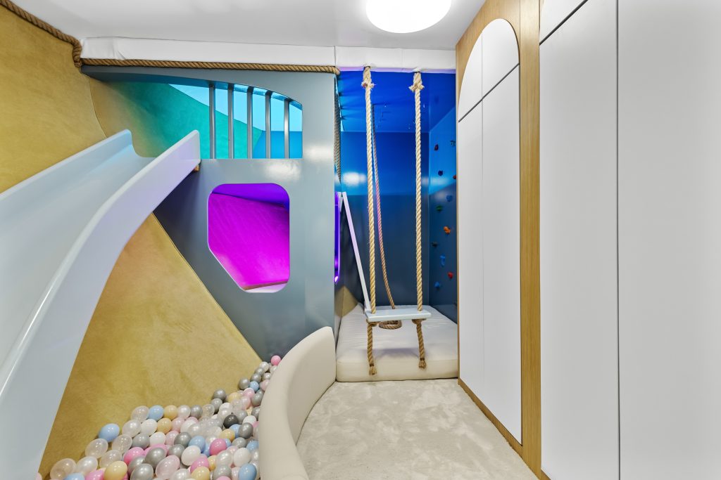 A modern indoor playroom with a ball pit, light blue slide, climbing wall, rope swing, and colorful ambient lighting in the background. The room has soft flooring and built-in storage cabinets.