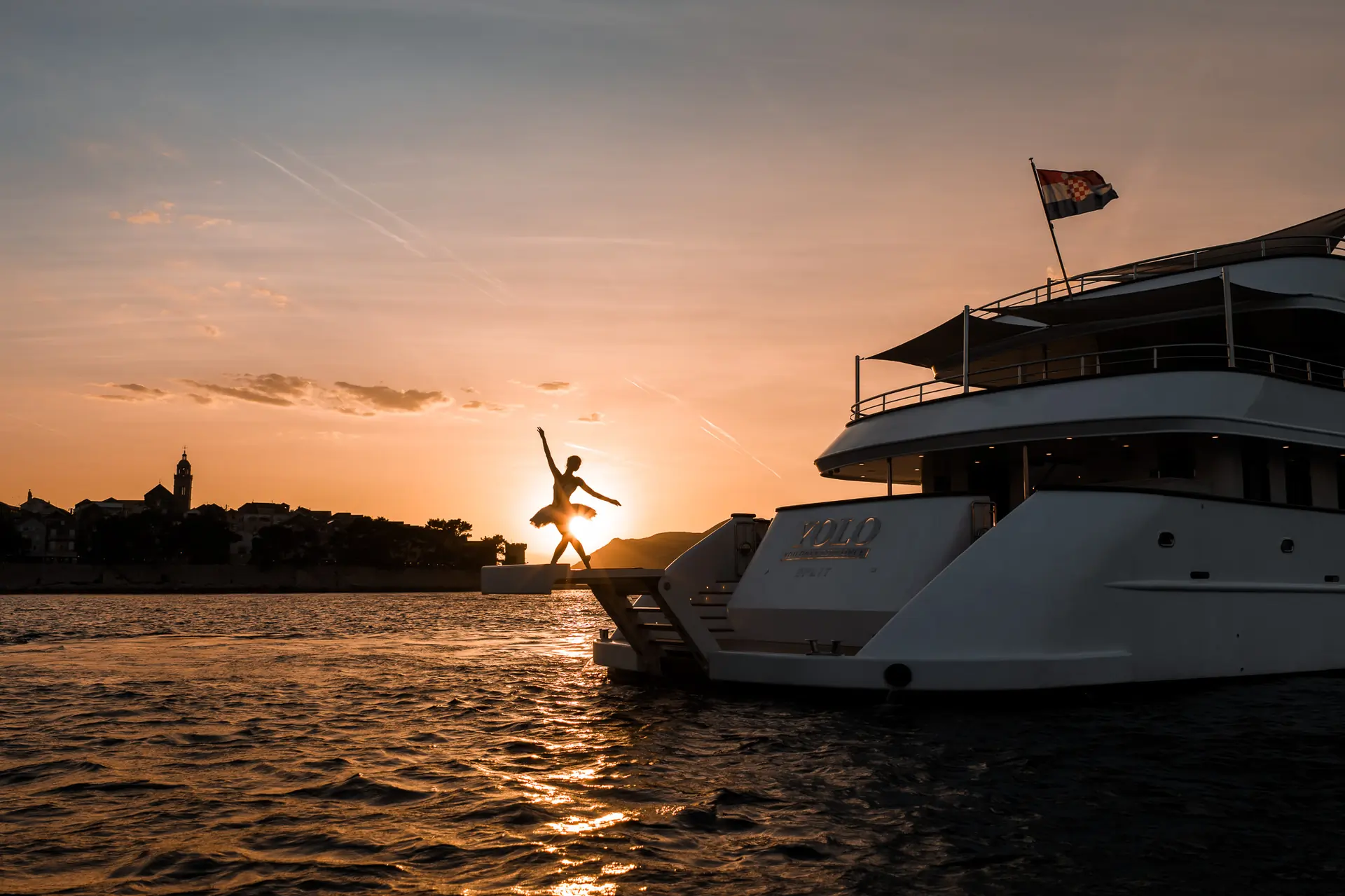 A person jumps off the back of a yacht into the water at sunset, with a city skyline and a flag visible in the background.