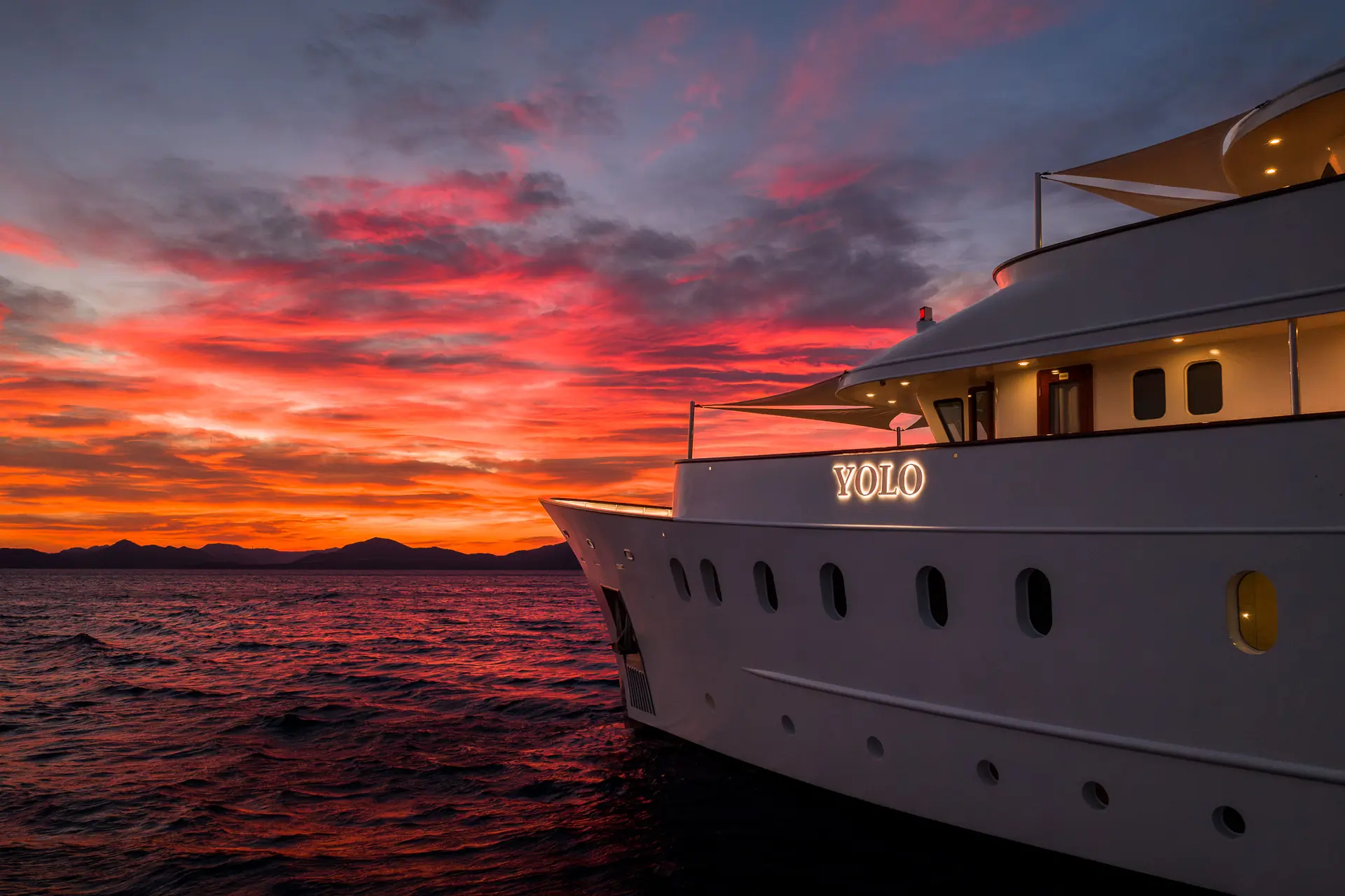 A white yacht named YOLO is anchored on the ocean at sunset, with vibrant red, orange, and purple clouds filling the sky and distant mountains visible on the horizon.