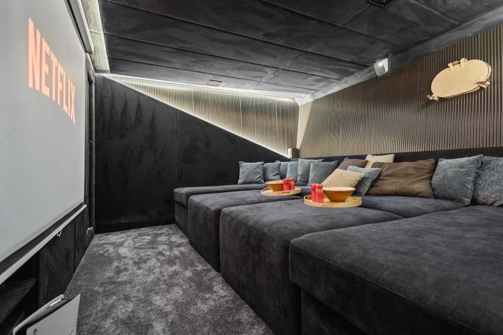 A cozy home theater room with dark walls, a large sectional sofa with pillows, two gold trays with snacks and drinks, and Netflix projected on a screen. The room has soft lighting and a modern, minimalist design.