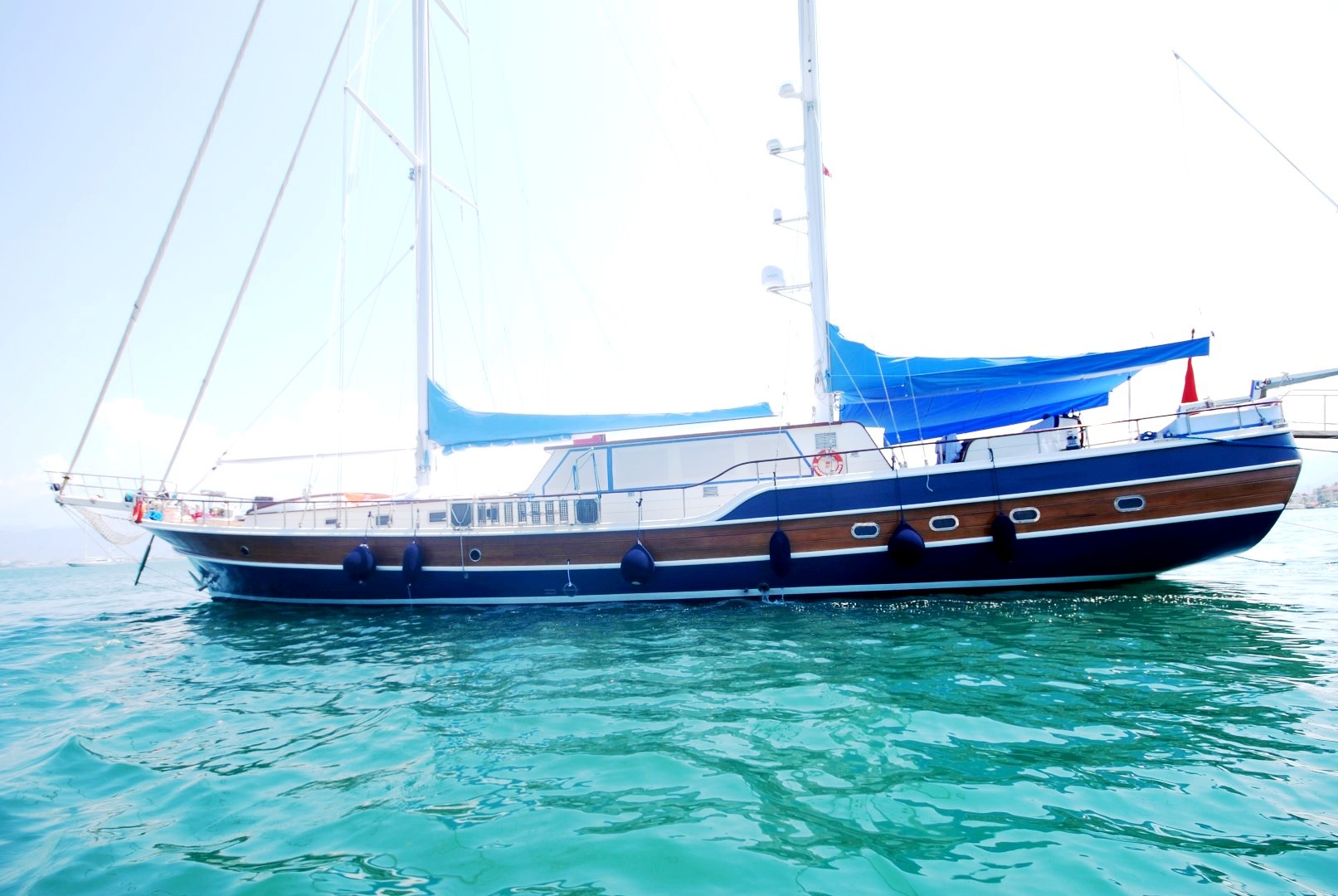 Un gran yate de madera con toldos azules está anclado en aguas tranquilas de color turquesa bajo un cielo brillante y despejado.