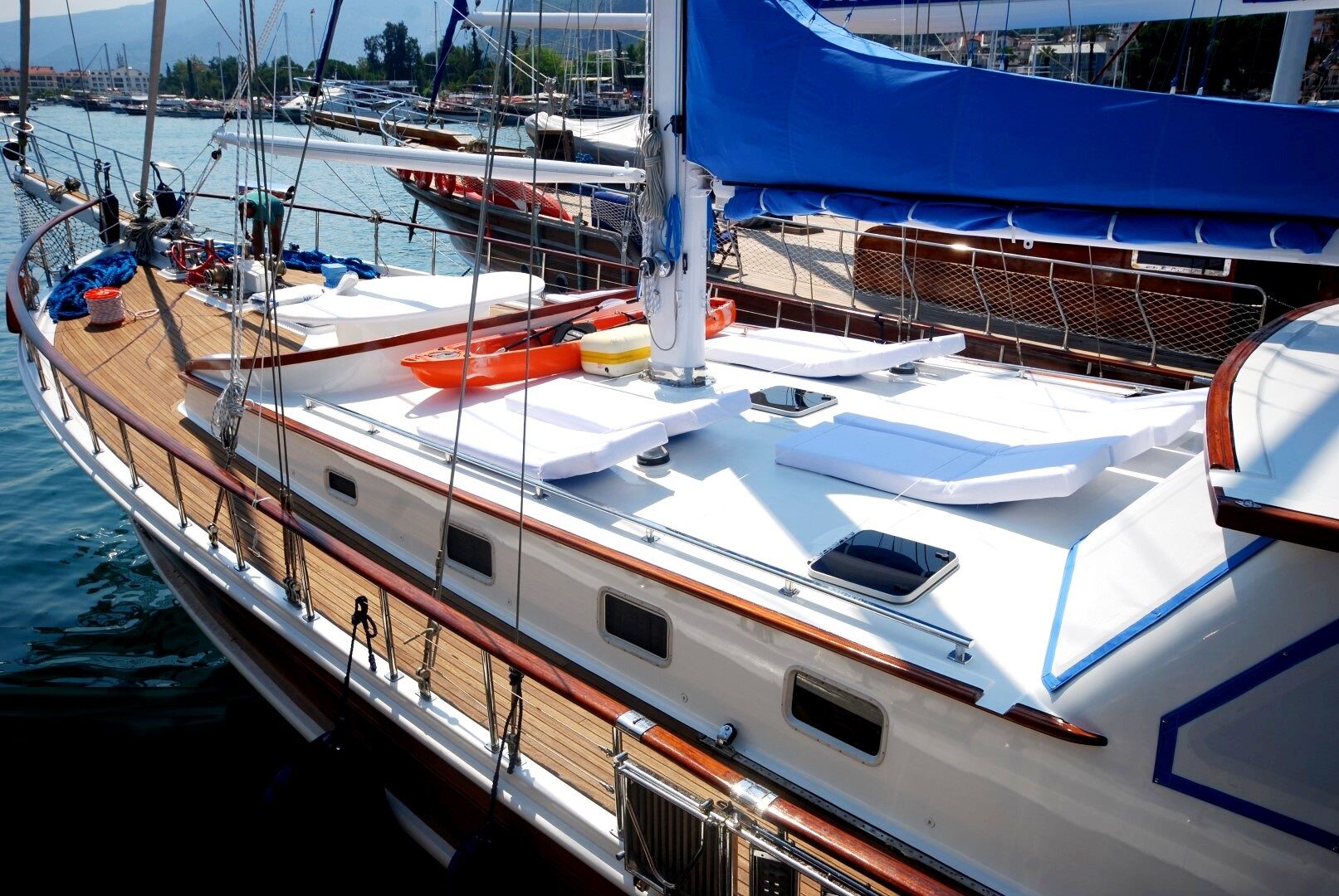 Un velero blanco con cubierta de madera está atracado en un puerto deportivo. El barco tiene cubiertas azules, cojines de salón blancos y aros salvavidas naranjas. Al fondo se ven otros barcos y un paseo marítimo.