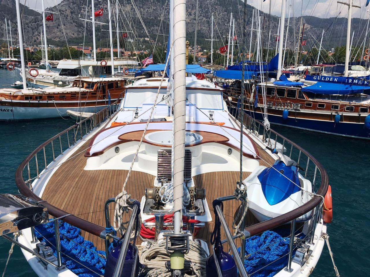 Vista desde la proa de un velero atracado en un puerto deportivo, con cubierta de madera, cuerdas enrolladas y otros barcos amarrados cerca; al fondo se ven montañas y banderas turcas.