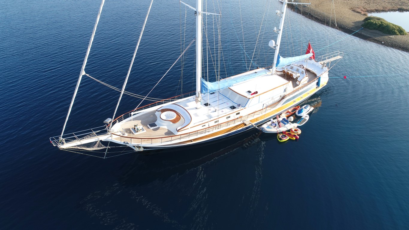 Vista aérea de un gran velero anclado en aguas tranquilas y azules cerca de la costa, con una pequeña lancha motora y varios flotadores hinchables amarrados al costado.