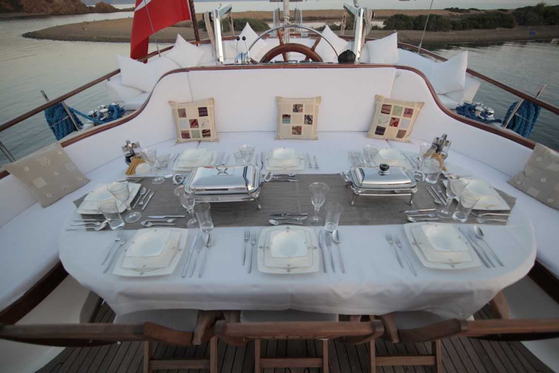 Una elegante mesa de comedor al aire libre para seis personas con mantelería blanca, cubertería de plata y vajilla cubierta en la cubierta de un barco. Asientos acolchados y cojines rodean la mesa, con el agua y la costa visibles al fondo.
