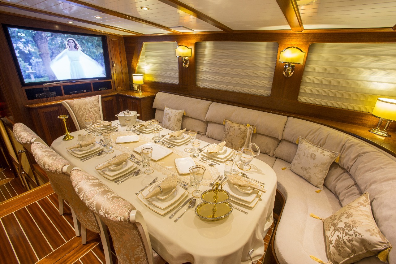 El lujoso comedor de un yate cuenta con una elegante mesa para ocho comensales, asientos de felpa, decoración ornamental, iluminación cálida y un gran televisor en el que aparece una mujer vestida de blanco.