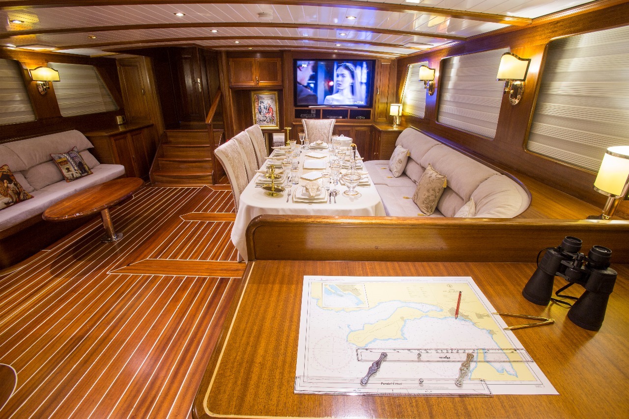 Lujoso interior de yate con suelos de madera, una mesa de comedor preparada para una comida, acogedores sofás, lámparas de pared, un televisor y una mesa de navegación con un mapa, un lápiz, una regla y unos prismáticos en primer plano.