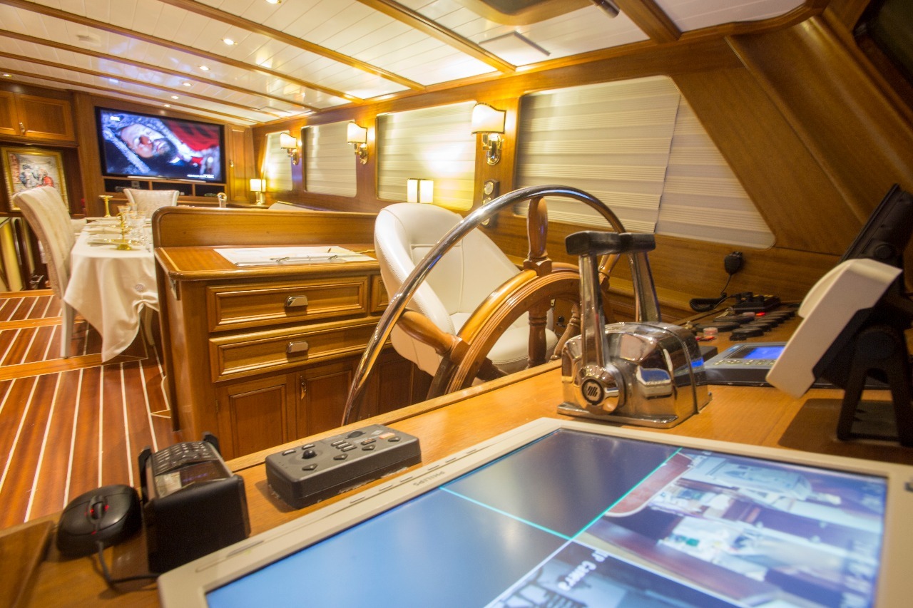 Interior de un lujoso yate con un volante de madera, controles de navegación y monitores en primer plano. Al fondo, una mesa de comedor con platos y vasos, y una gran pantalla en la pared.