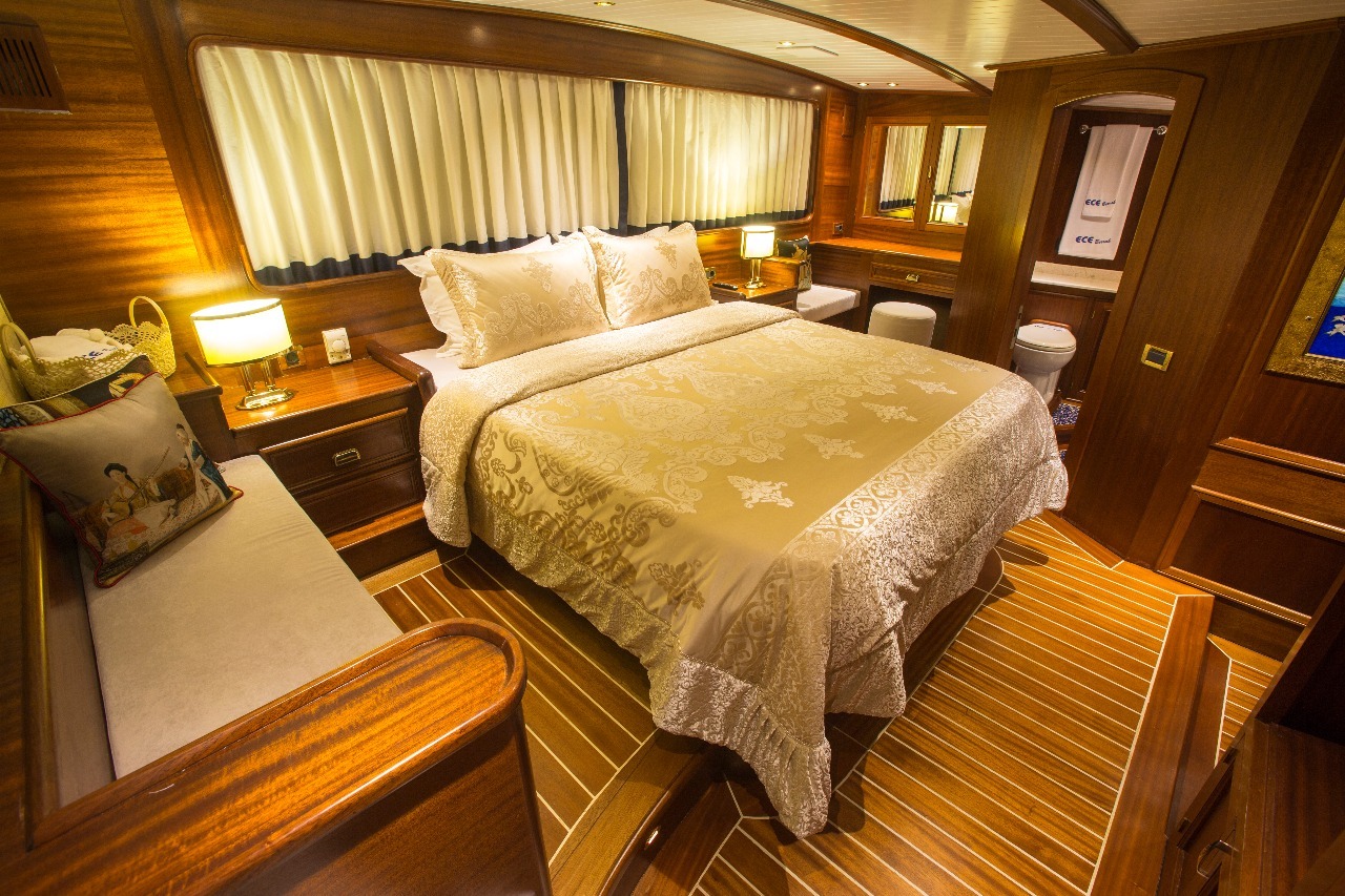 Lujoso camarote de barco con una gran cama cubierta con ropa de cama de color crema ornamentada, paredes y suelos de madera, iluminación tenue, un banco acolchado, espejos y toallas cuidadosamente dispuestas en una estantería en un rincón.