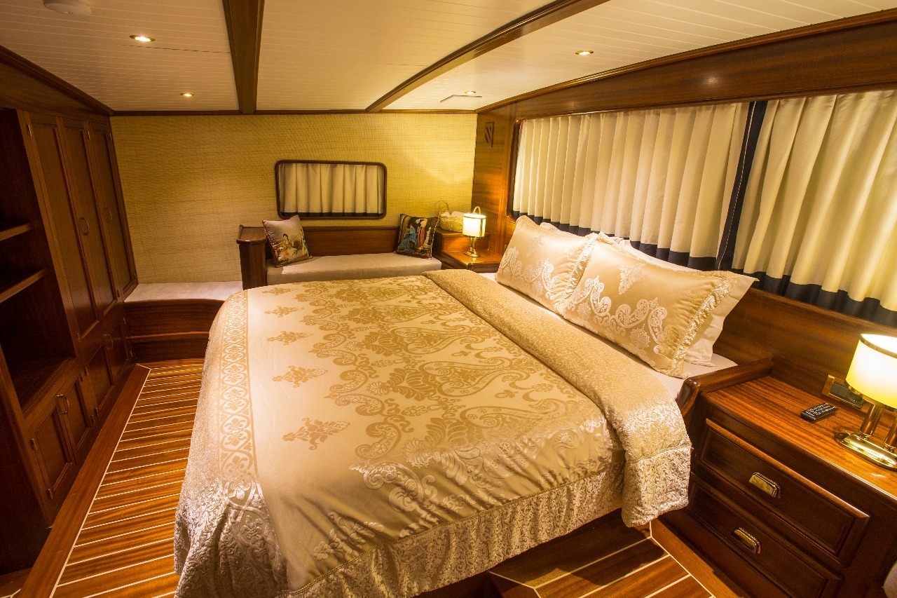 Lujoso dormitorio en camarote con una gran cama con motivos dorados, suelos y paredes de madera, estanterías empotradas, mesillas de noche con lámparas, cortinas sobre las ventanas y un banco acolchado con almohadas decorativas.