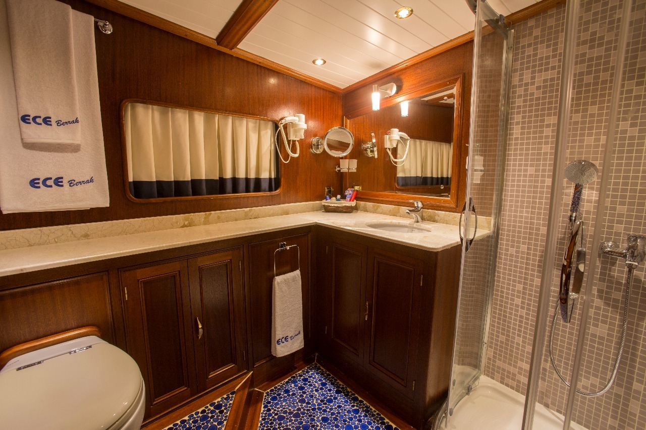 Un lujoso cuarto de baño de yate con armarios de madera, encimera de mármol, un gran espejo, ducha de cristal, toallas con la etiqueta ECCE Berrak y baldosas de mosaico azul. El espacio está bien iluminado y organizado.