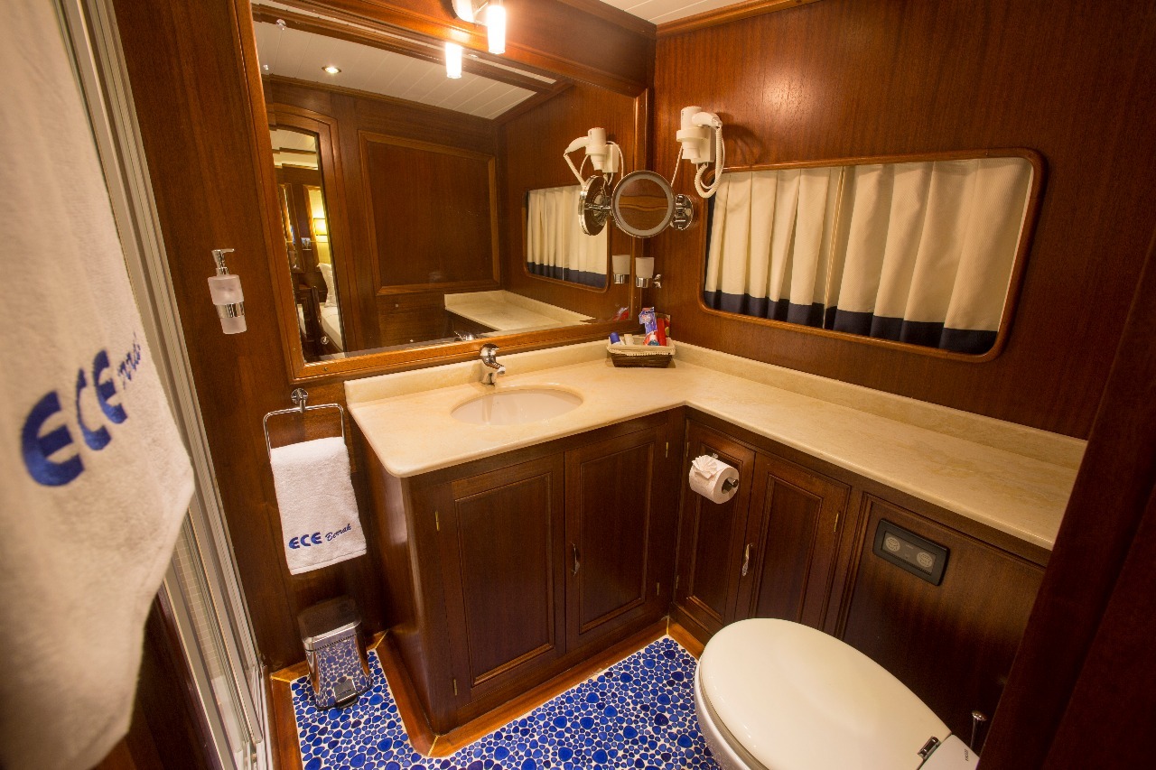 Un cuarto de baño con armarios de madera, un inodoro blanco, un lavabo con encimera de mármol, un espejo de pared, toalleros con toallas blancas y suelo de baldosas con motivos azules. En el mostrador hay un secador de pelo y artículos de aseo.