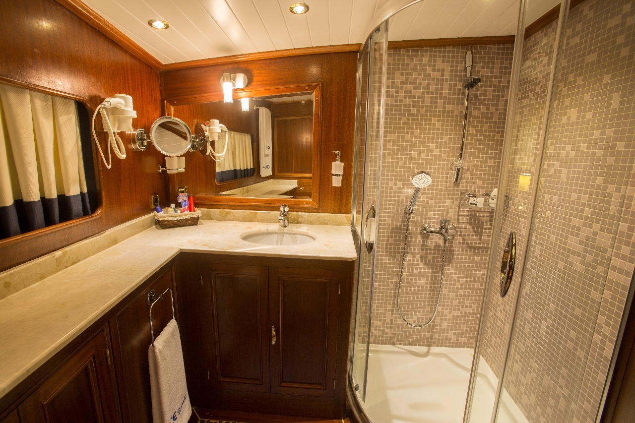 Un cuarto de baño bien iluminado con armarios de madera, una encimera de mármol, un lavabo, un espejo grande, un secador de pelo montado y una ducha acristalada con cabezal de ducha de lluvia y de mano, además de paredes de azulejos de mosaico.