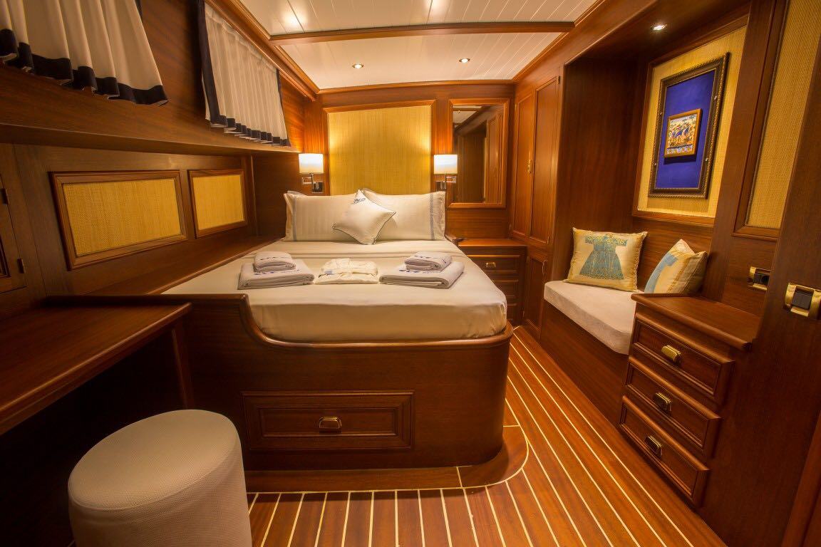 Lujoso camarote de yate con paneles de madera, una cama doble bien hecha, toallas dobladas, un banco acolchado con cojines, obras de arte enmarcadas en la pared y una iluminación cálida que crean un ambiente acogedor y elegante.