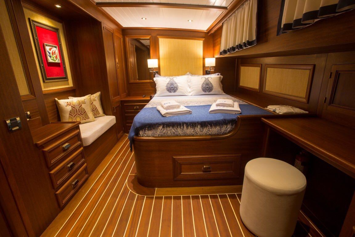 Un acogedor camarote de yate con interior de madera, que cuenta con una cama doble con ropa de cama blanca y azul, almohadas, un banco con cojines, cajones, zona de escritorio y la suave iluminación de dos lámparas de cabecera.