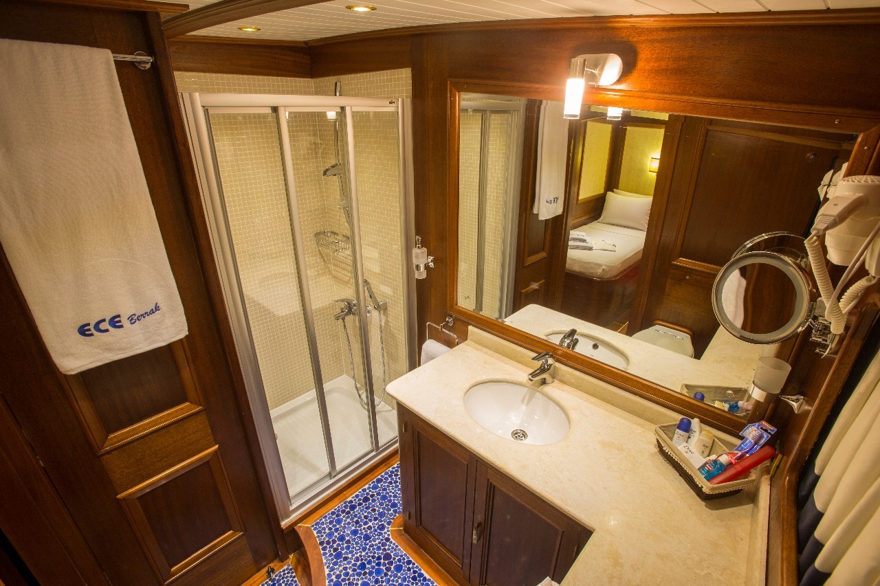 Un pequeño cuarto de baño con armarios de madera, un lavabo con encimera de mármol, una cabina de ducha de cristal, grandes espejos, una toalla montada en la pared, baldosas de mosaico azul en el suelo y artículos de aseo ordenados junto al lavabo.