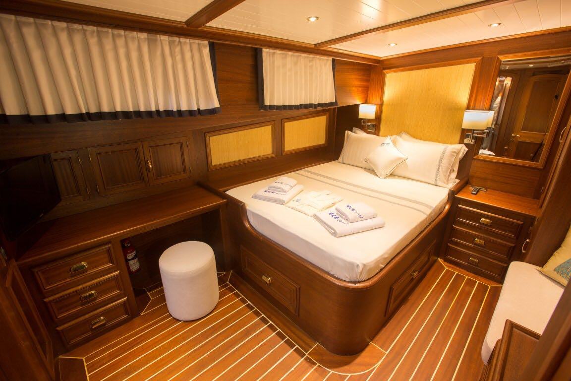 Un acogedor camarote de yate con paredes y suelo revestidos de madera, una cama doble bien hecha, lámparas junto a la cama, un escritorio empotrado con un taburete blanco y toallas dobladas sobre la cama. El espacio parece cálido y acogedor.