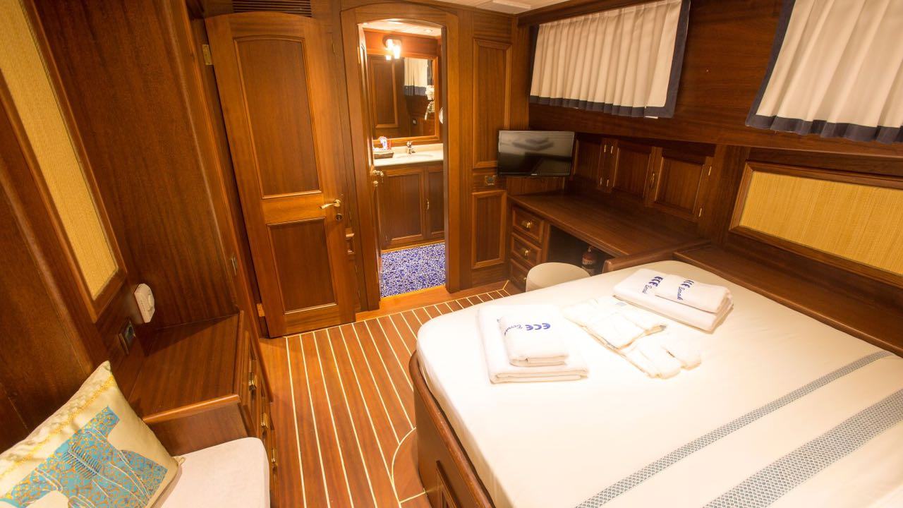 Un acogedor camarote con paredes revestidas de madera, una cama doble con toallas dobladas, un escritorio con una silla, un televisor de pantalla plana y una puerta que da a un cuarto de baño con lavabo y espejo. Cortinas blancas cubren las ventanas.