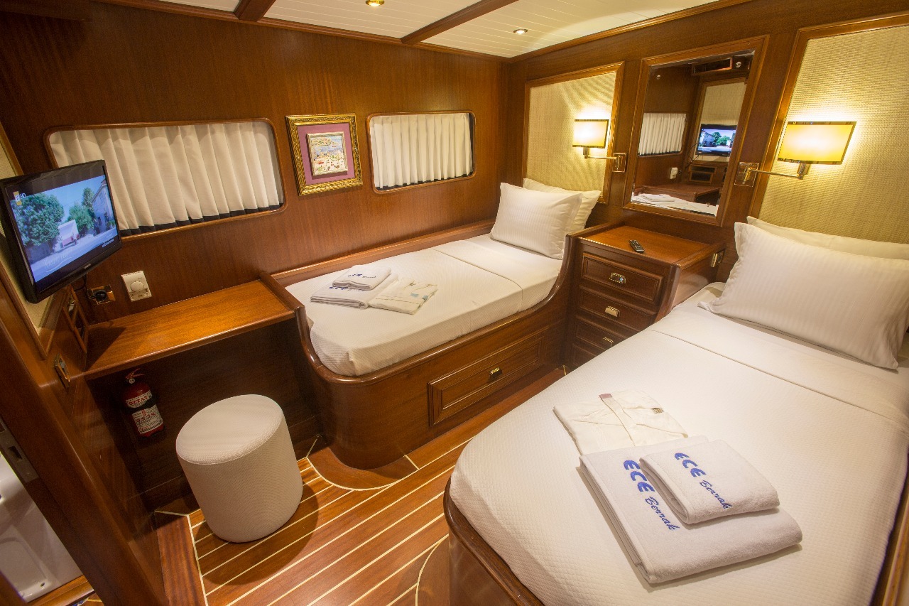 Un acogedor camarote de yate con paredes de madera pulida, dos camas individuales, un pequeño escritorio, un televisor montado en la pared, un espejo, iluminación tenue y toallas cuidadosamente dobladas en cada cama.