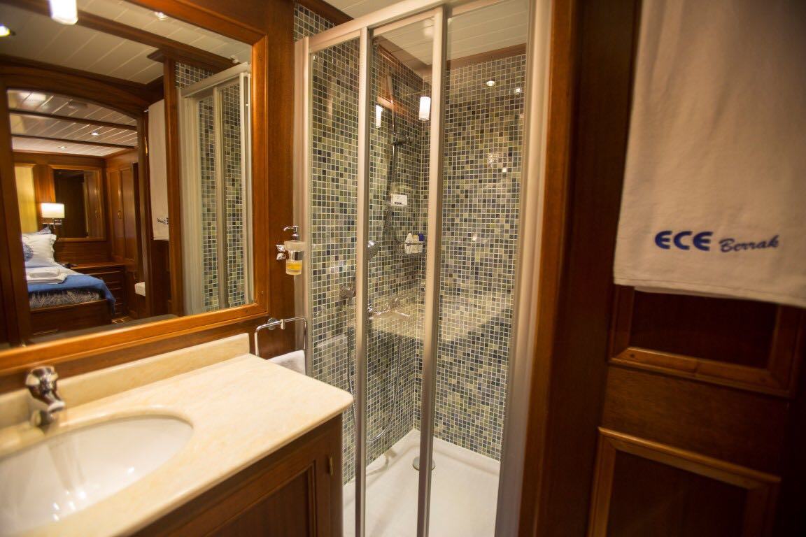 Un cuarto de baño compacto con una ducha acristalada, paredes de azulejos de mosaico, un lavabo blanco con encimera de mármol, un espejo y una toalla colgada de una puerta de madera. En la habitación contigua se ve una cama.