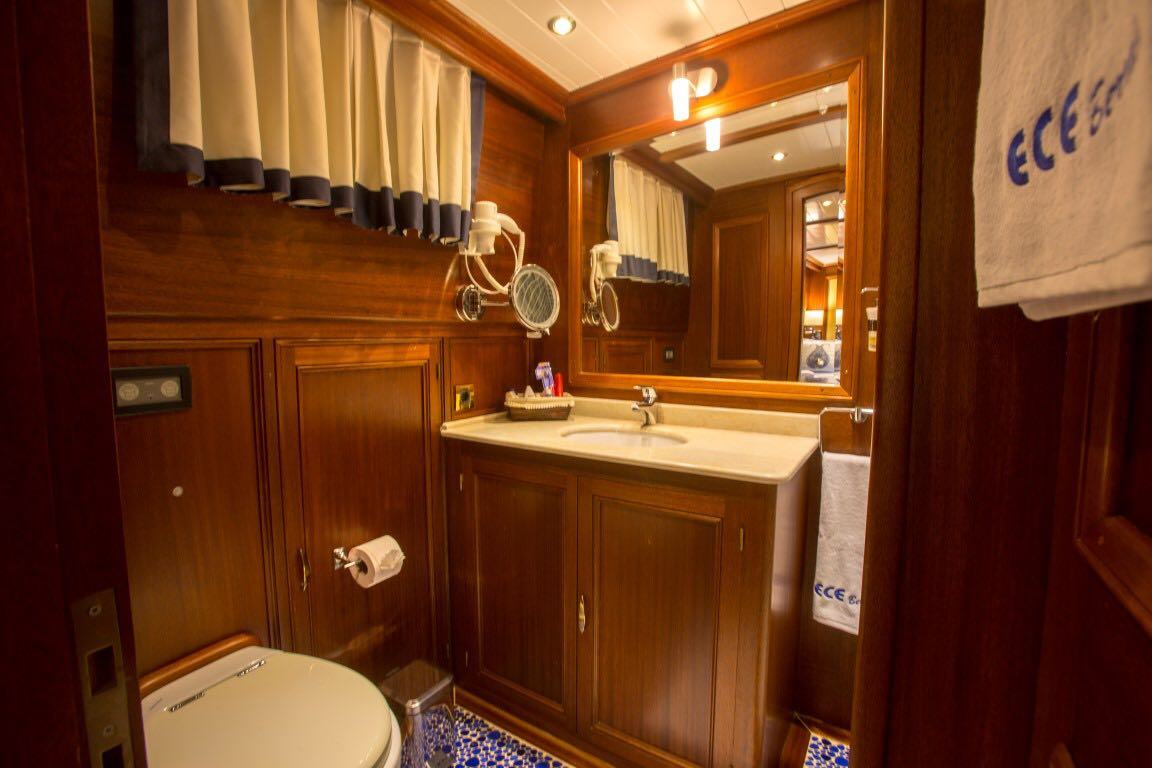 Un baño acogedor con paredes y armarios de madera, un lavabo con un gran espejo, un secador de pelo montado en la pared, un inodoro, suelo de baldosas con motivos azules y toallas colgadas en la pared. La ventana tiene cortinas de color crema y azul marino.