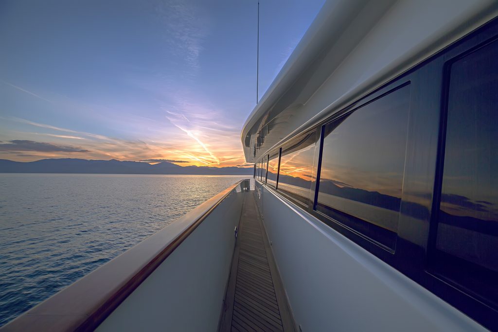 View from the side deck of a yacht charter at sunset, showing the calm sea, distant mountains, and colorful sky reflected in the yacht’s windows.