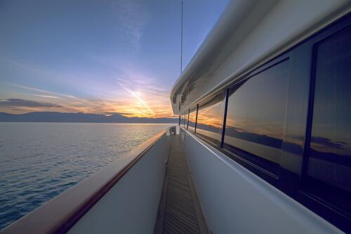 View from the side deck of a yacht charter at sunset, showing the calm sea, distant mountains, and colorful sky reflected in the yacht’s windows.